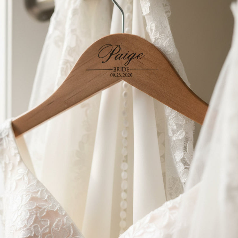 Wooden hanger with 'Paige BRIDE 09.23.2026' engraved on a white lace wedding dress.