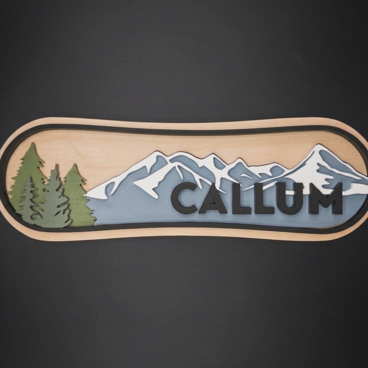 Decorative patch shaped like a snowboard with mountains, trees, and the name 'Callum' on a dark background